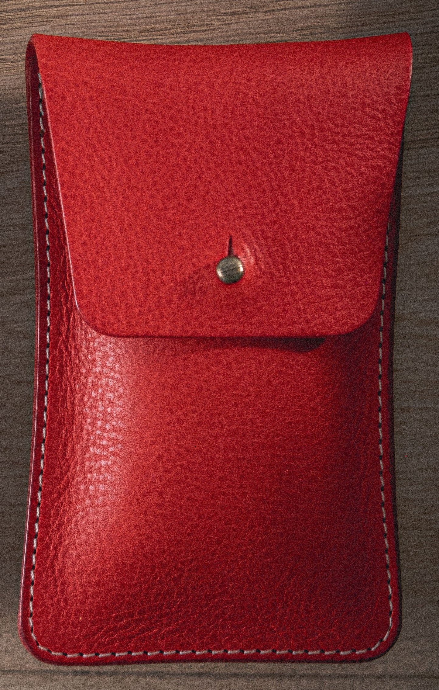 A vibrant red pebbled veg-tan leather watchpouch from the "Vibrant" collection, featuring a flap secured by a single, small metallic button and highlighted with white stitching along the edges. Placed on a wooden surface, it beautifully showcases its handmade craftsmanship from Antwerp.