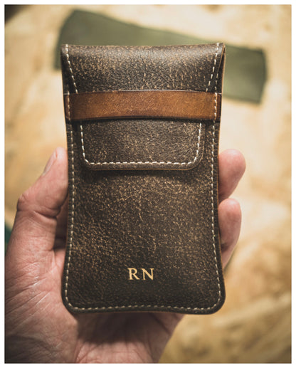 A hand holds a premium leather pouch from the Soft Layered Premium Pouch Collection (with lining), handmade in Antwerp. The brown leather case, featuring white stitching along the edges, has a front flap secured by a horizontal strap and is embossed with the initials "RN" in gold at the bottom. The background is a blurred wooden surface.