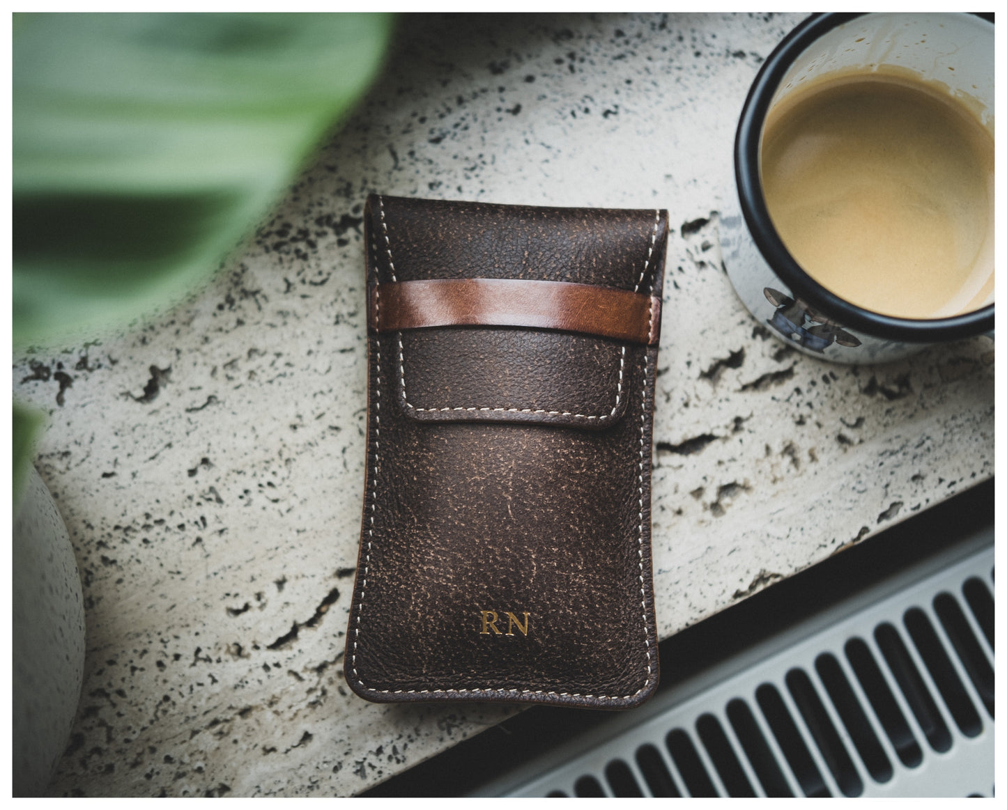 A brown, textured leather item from the Soft Layered Premium Pouch Collection (with lining) with stitching and a small strap lies next to a cup of creamy coffee on a speckled surface. The initials "R.N" are embossed in gold near the bottom of this premium watch accessory. A green plant is partially visible in the top left corner.