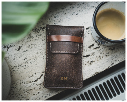 A brown, textured leather item from the Soft Layered Premium Pouch Collection (with lining) with stitching and a small strap lies next to a cup of creamy coffee on a speckled surface. The initials "R.N" are embossed in gold near the bottom of this premium watch accessory. A green plant is partially visible in the top left corner.