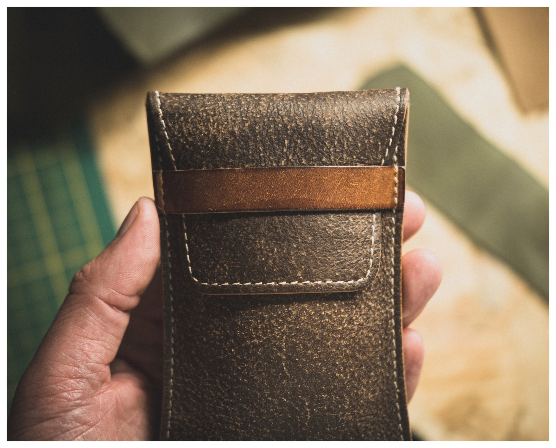 Close-up of a hand holding a Soft Layered Premium Pouch with visible stitching and a flap closure. The background is softly focused, revealing a cutting mat and other leather pieces, suggesting a workshop setting where this high-quality watch accessory from the Soft Layered Premium Pouch Collection (with lining) was handmade in Antwerp.