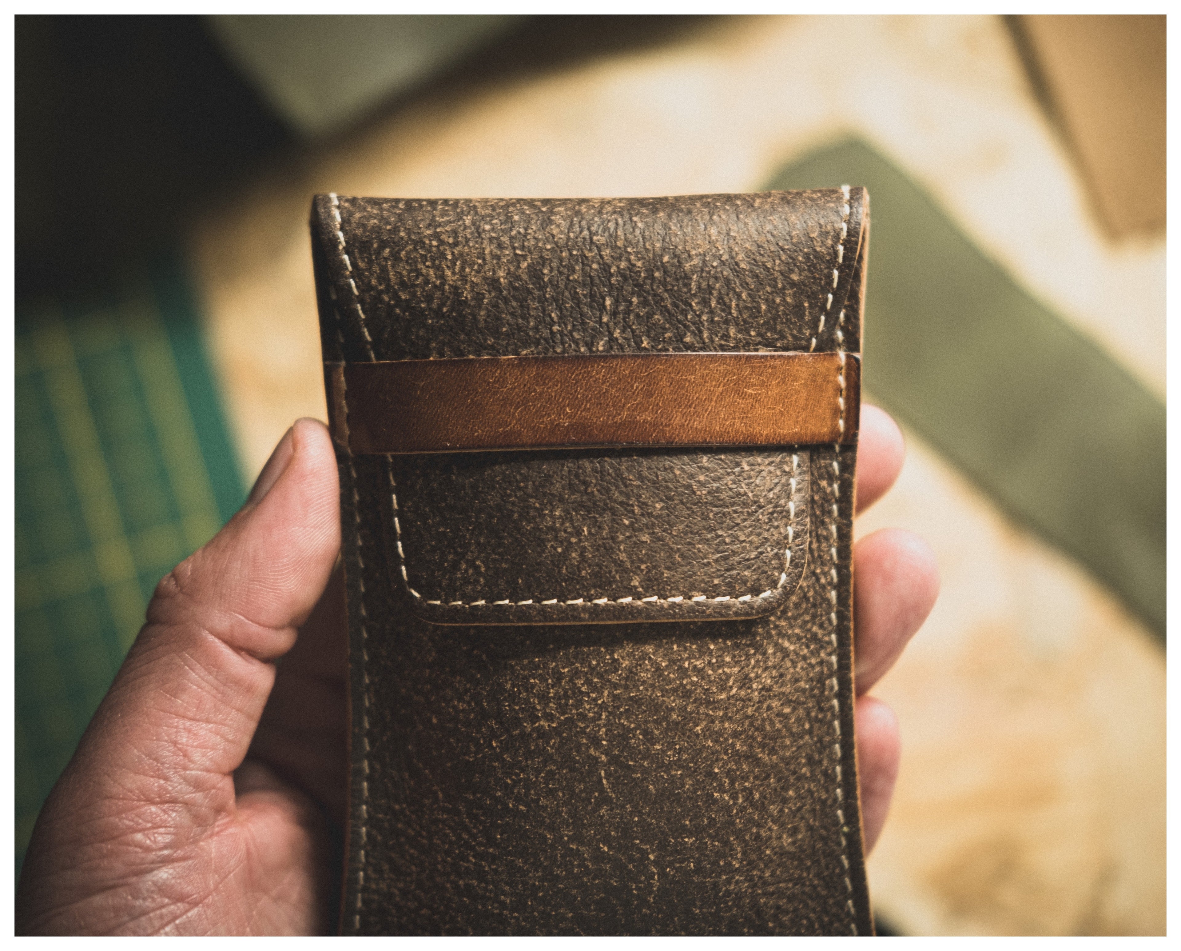 Close-up of a hand holding a Soft Layered Premium Pouch with visible stitching and a flap closure. The background is softly focused, revealing a cutting mat and other leather pieces, suggesting a workshop setting where this high-quality watch accessory from the Soft Layered Premium Pouch Collection (with lining) was handmade in Antwerp.