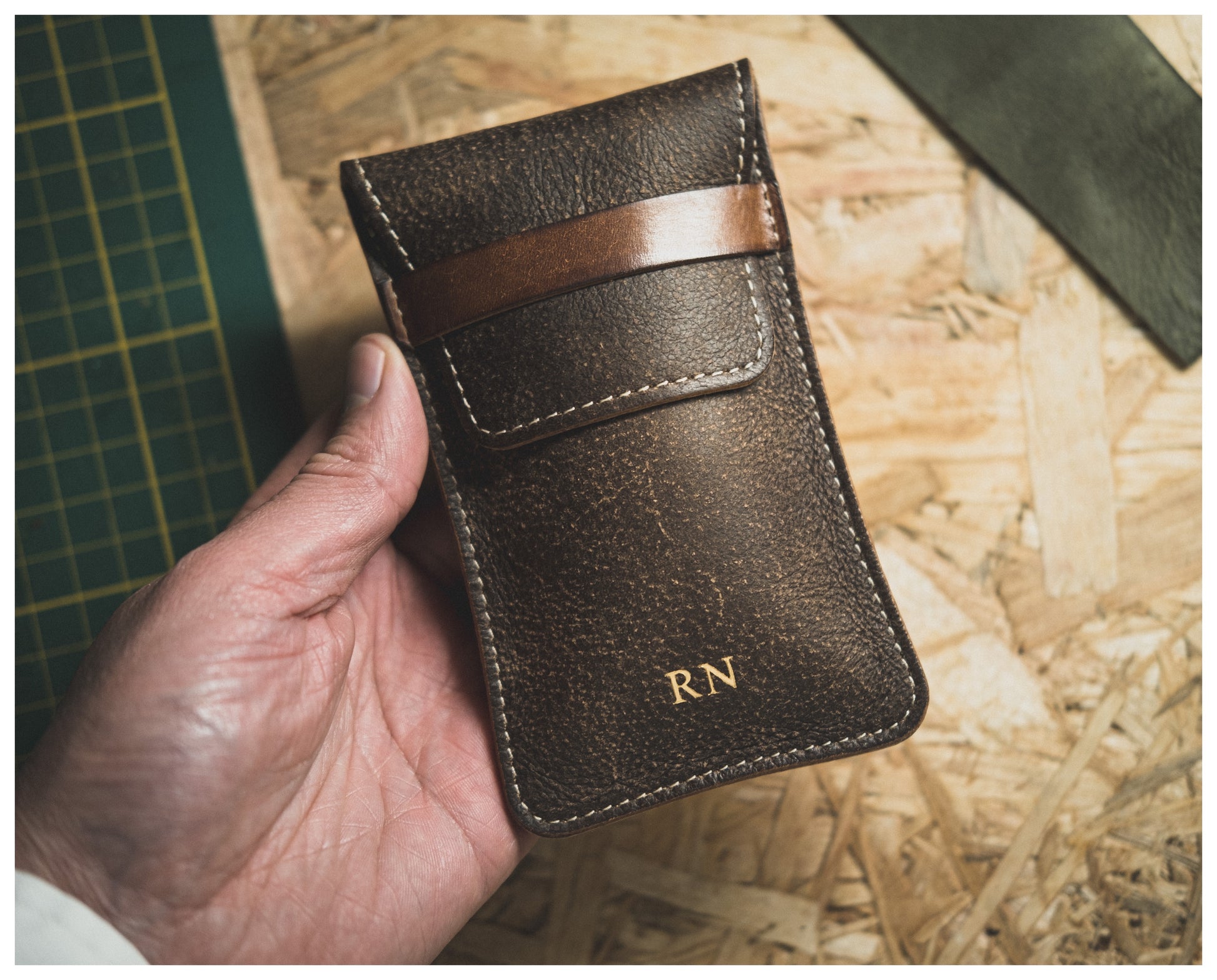 A hand is holding a stitched dark brown leather pouch from the Soft Layered Premium Pouch Collection (with lining), featuring a horizontal strap and the initials "RN" embossed in gold at the bottom. This luxurious watch accessory, handmade in Antwerp, rests against a green cutting mat and a textured wood surface.