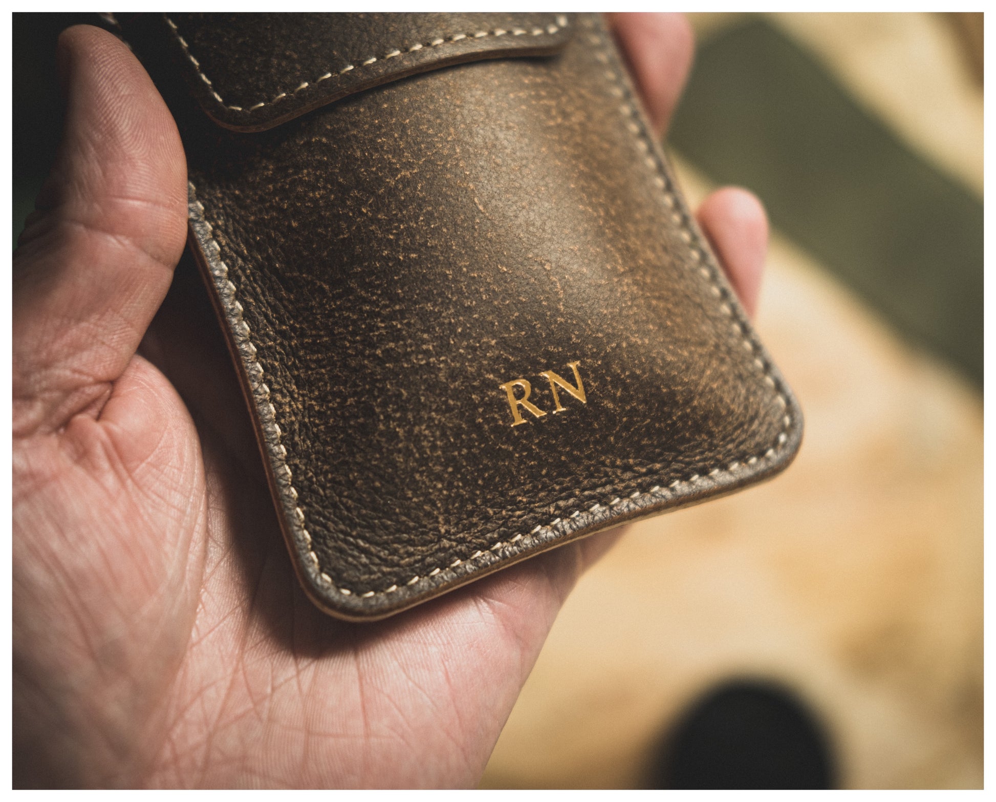 A hand holding a small, brown leather watch pouch from the Soft Layered Premium Pouch Collection, with the initials "RN" embossed in gold letters on the surface. Handmade in Antwerp, this elegant watch accessory rests against a slightly out-of-focus background of wooden surface and green cloth.
