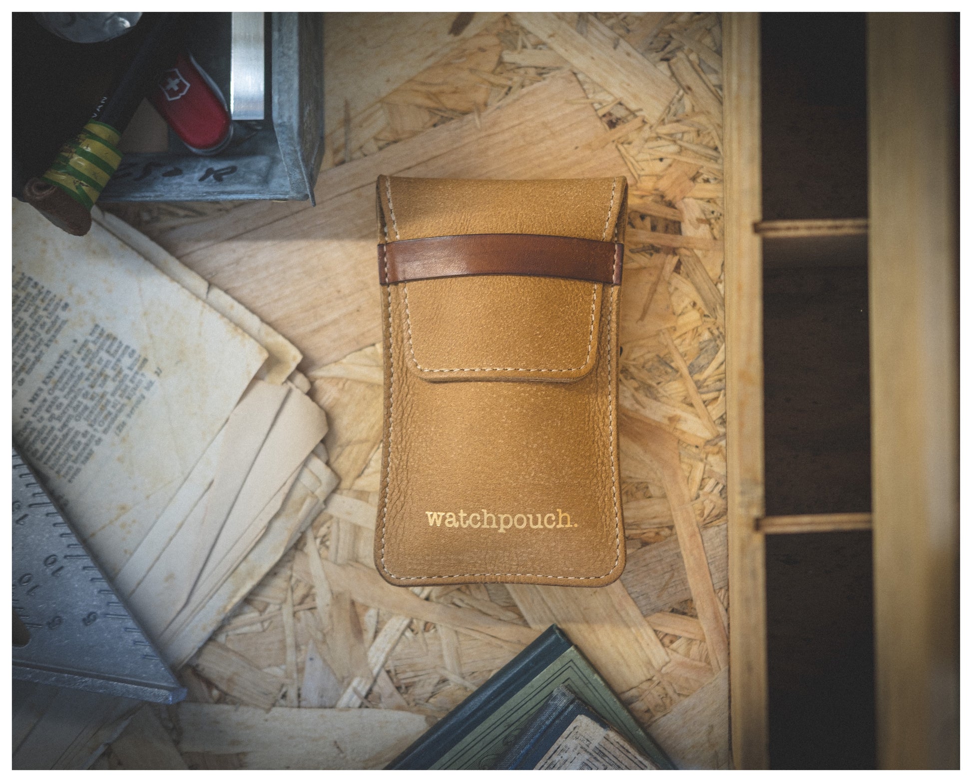 A tan leather pouch from the Soft Layered Premium Pouch Collection, embossed with the word "watchpouch.", is placed on a wooden surface surrounded by vintage paper documents, a partially visible closed book, and a red pocket knife. This premium watch accessory, handmade in Antwerp, adds to the scene's rustic, nostalgic feel.
