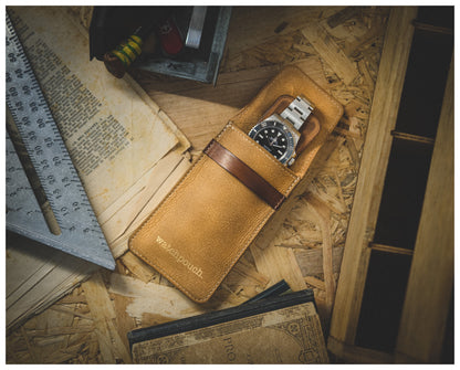 A wristwatch with a metal band is housed in a tan, rectangular Soft Layered Premium Pouch Collection (with lining) placed on a wooden surface. The surface is scattered with old papers, a square metal ruler, and other vintage items, creating a rustic vibe. This premium watch accessory was handmade in Antwerp.