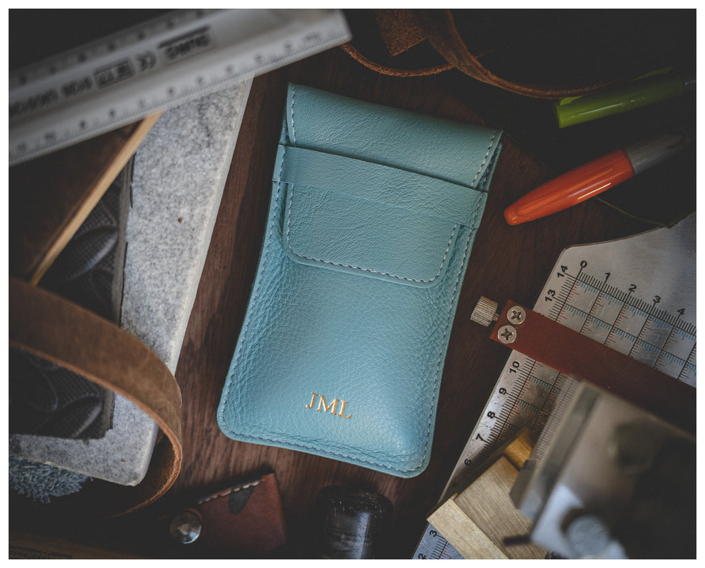 A light blue leather watch pouch from the Soft Layered Premium Pouch Collection, adorned with gold initials "JML" on the front, is surrounded by various tools including a ruler, protractor, red and green pens, and leather strips. This premium handmade watch accessory from Antwerp is elegantly displayed on a wooden surface.