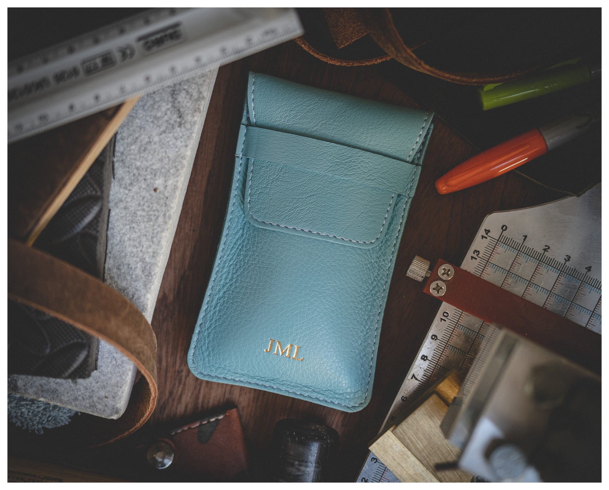 A light blue leather watch pouch from the Soft Layered Premium Pouch Collection, adorned with gold initials "JML" on the front, is surrounded by various tools including a ruler, protractor, red and green pens, and leather strips. This premium handmade watch accessory from Antwerp is elegantly displayed on a wooden surface.