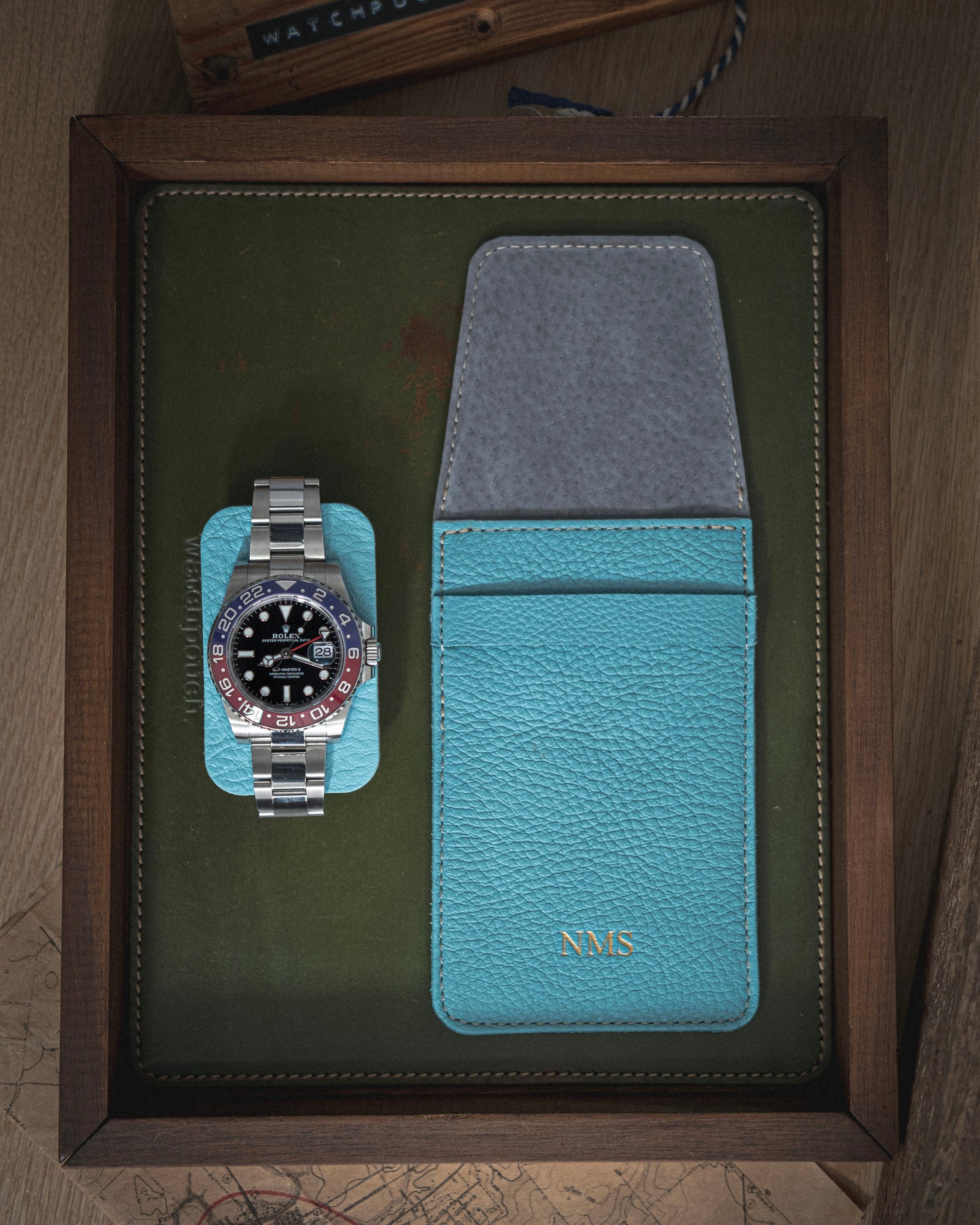 A luxury wristwatch featuring a silver band, red and blue bezel, and black dial is displayed on a light blue case. Next to it is a handmade premium leather watch pouch from the Soft Layered Premium Pouch Collection with initials "NMS," crafted in Antwerp. Both items are set within a dark wooden frame accented by green and gray background elements.