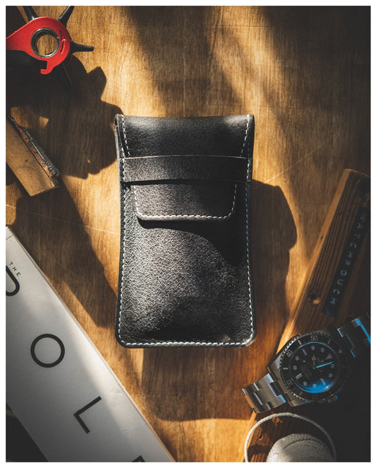 A soft layered premium pouch from the collection, featuring white stitching, is centered on a wooden surface. Surrounding it are various items, including a leather watch accessory, a harmonica, a book, and a small red tool. Sunlight casts shadows across the scene, creating a warm and rustic ambiance perfect for this handmade item from Antwerp.