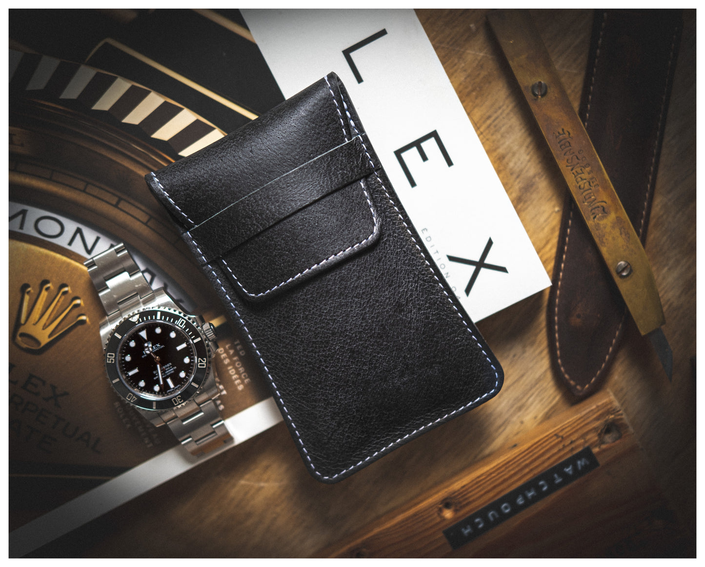 A flat-lay photo displaying a black leather wallet with white stitching, a silver wristwatch with a black face, a wooden-handled tool, a handmade leather watch pouch from Antwerp named "Soft layered premium pouch collection (with lining)," and a book featuring the brand name "Rolex." The premium watch accessories are arranged on a wooden surface.