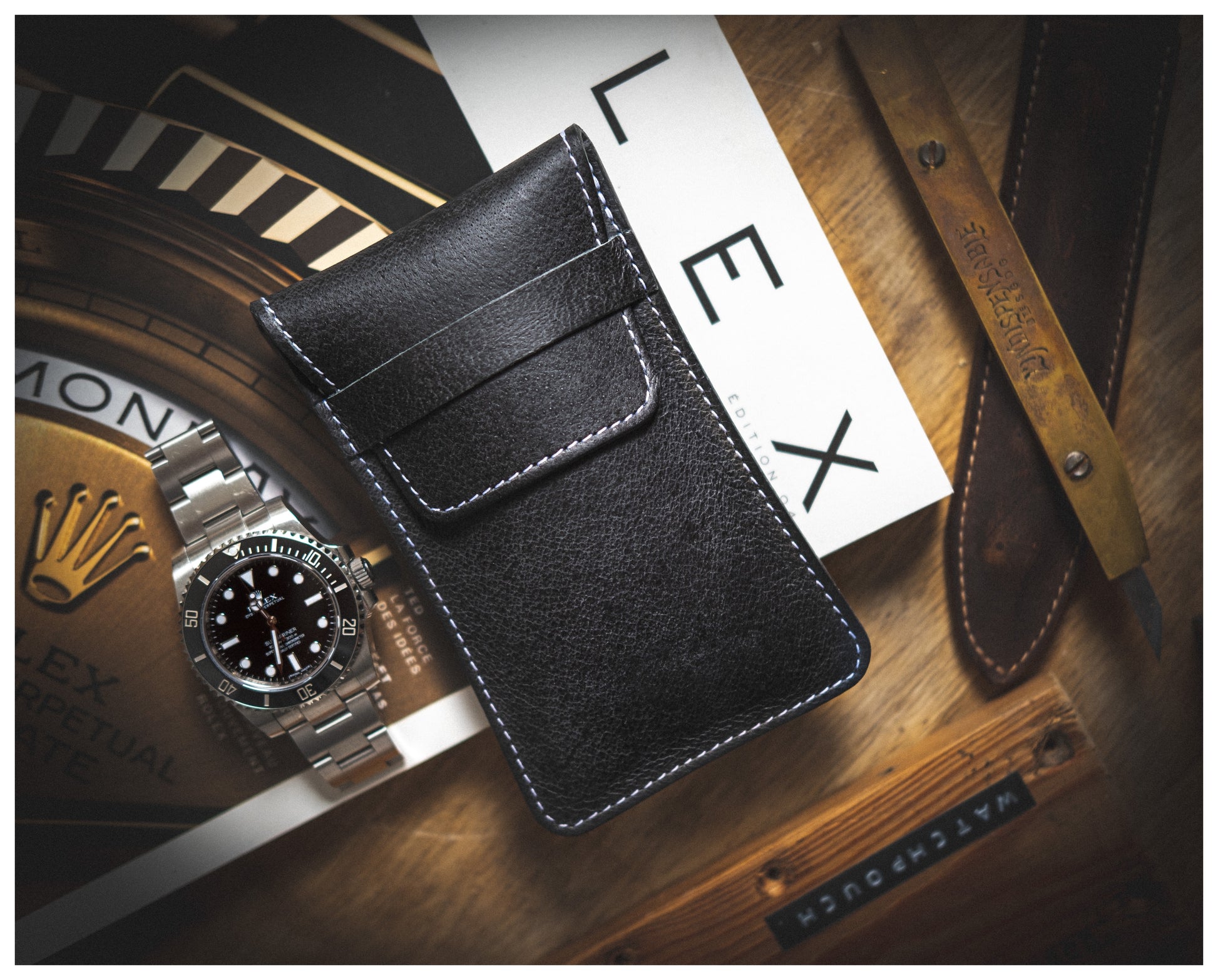 A flat-lay photo displaying a black leather wallet with white stitching, a silver wristwatch with a black face, a wooden-handled tool, a handmade leather watch pouch from Antwerp named "Soft layered premium pouch collection (with lining)," and a book featuring the brand name "Rolex." The premium watch accessories are arranged on a wooden surface.