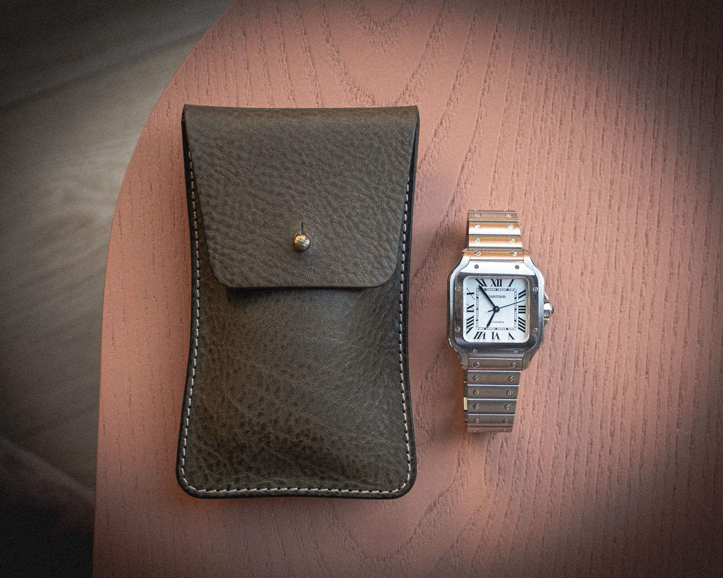 A stainless steel rectangular wristwatch with Roman numeral markings sits alongside a Pebbled veg-tan collection "Earth tones" leather pouch with a snap button, placed side by side on a wooden surface.