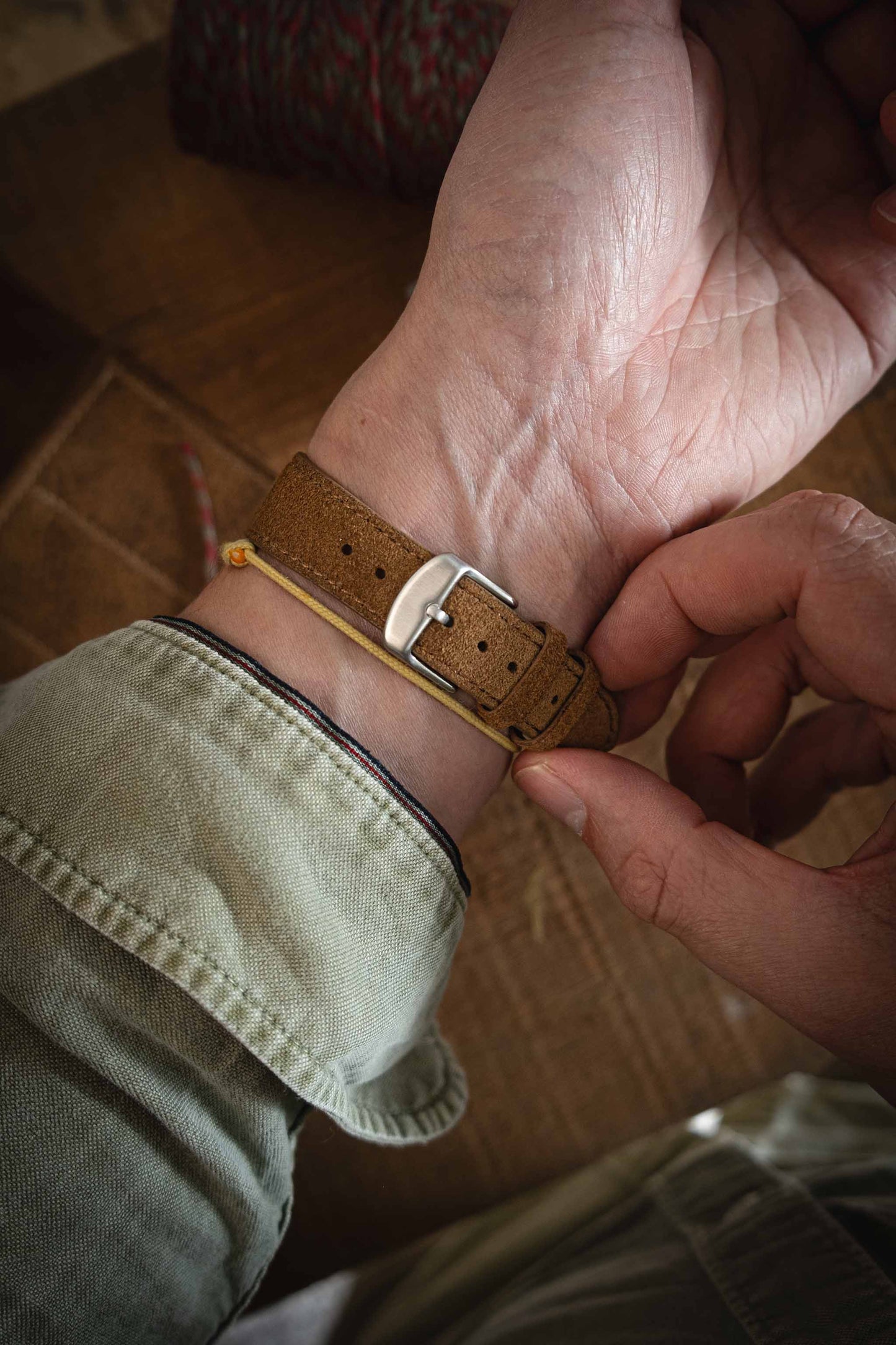 A person adjusts the brown suede watch strap on their wrist while wearing a light green shirt, with the focus on the watch buckle and strap.