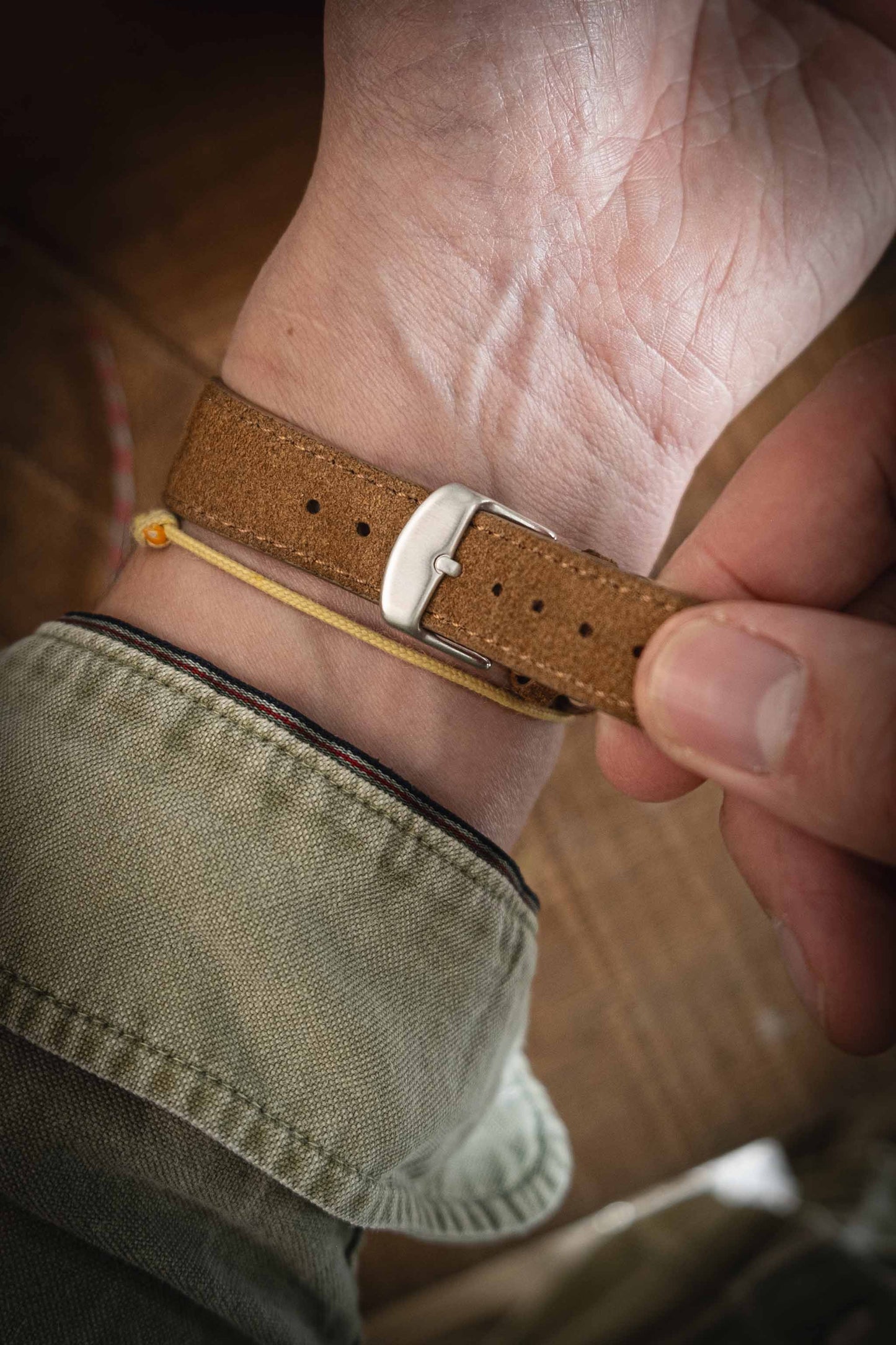 A close-up shows a person's wrist being fastened with a Suede watch strap in brown leather, featuring a quick-release spring bar. The person is wearing a green sleeve, and there's a yellow thread wrapped around the wrist beneath the strap.