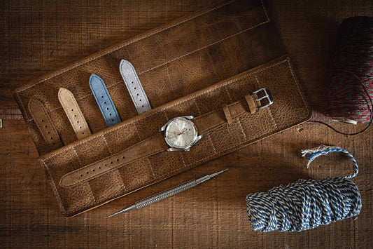 A stylish display features a watch on a brown leather case, surrounded by an assortment of Suede watch straps in various colors. On the wooden surface nearby, a metal tool and rolls of twine add to the scene, simplifying strap changes with the quick-release spring bar mechanism.