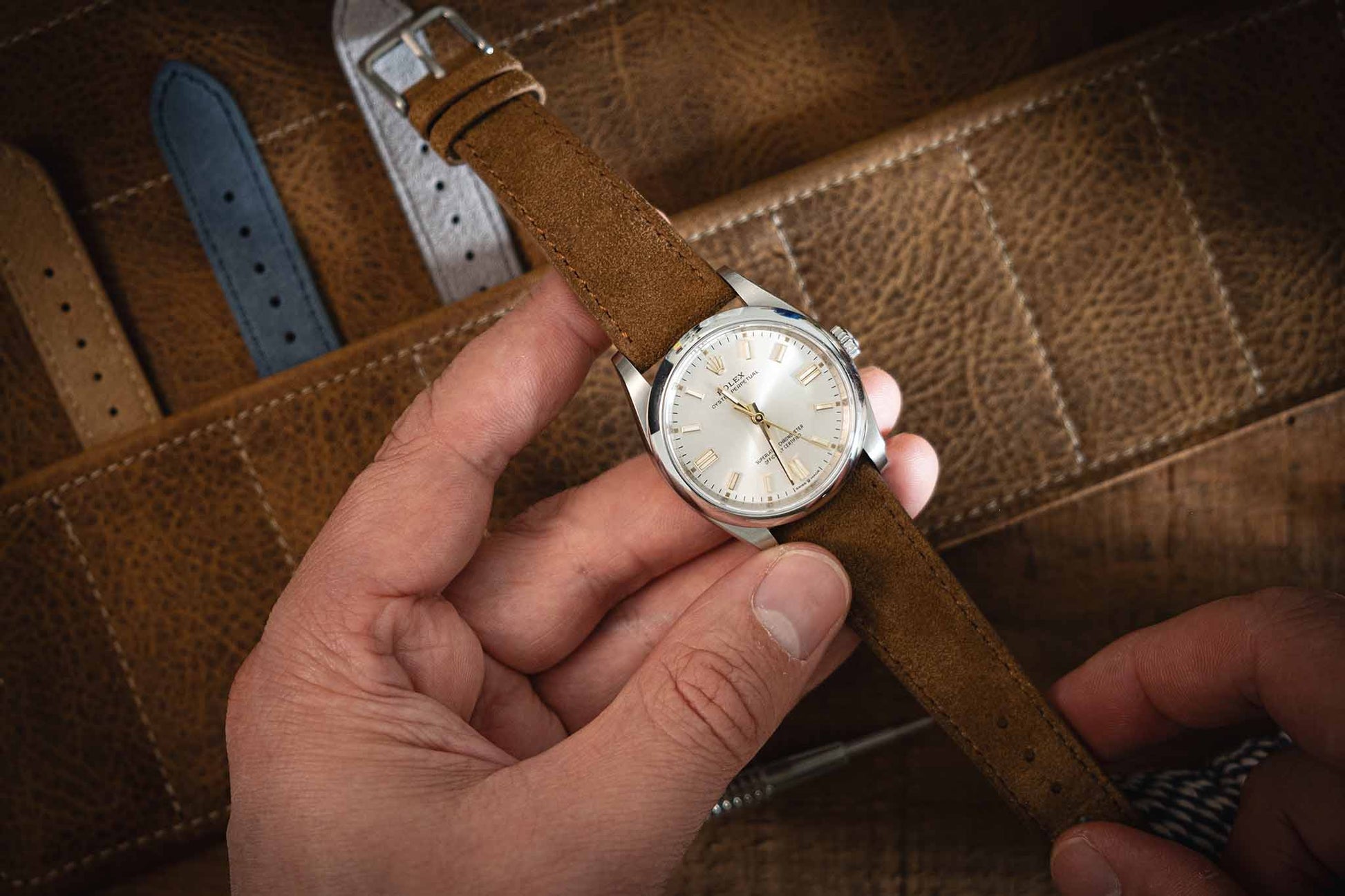 A person holds a wristwatch featuring one of the Suede watch straps in brown over a leather surface. The watch face is white with silver markers, and several additional straps, including handmade Suede watch straps, are displayed in the background.