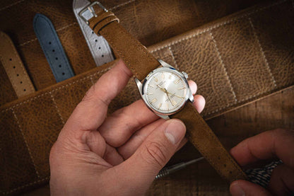 A person holds a wristwatch featuring one of the Suede watch straps in brown over a leather surface. The watch face is white with silver markers, and several additional straps, including handmade Suede watch straps, are displayed in the background.