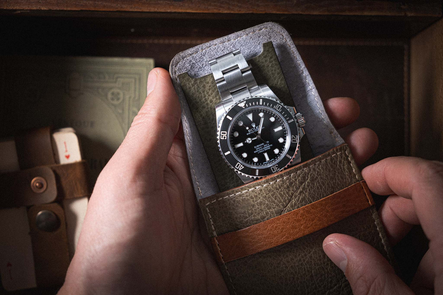 A person holds a stainless steel wristwatch with a black dial and bezel, nestled in a green leather case lined with premium leather. The watch boasts a metal bracelet and large, clear markings. In the background are visible a wooden box and an additional strap, along with a handmade pouch from Belgium from the Soft Layered Premium Pouch Collection featuring lining.