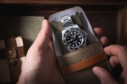 A person holds a stainless steel wristwatch with a black dial and bezel, nestled in a green leather case lined with premium leather. The watch boasts a metal bracelet and large, clear markings. In the background are visible a wooden box and an additional strap, along with a handmade pouch from Belgium from the Soft Layered Premium Pouch Collection featuring lining.