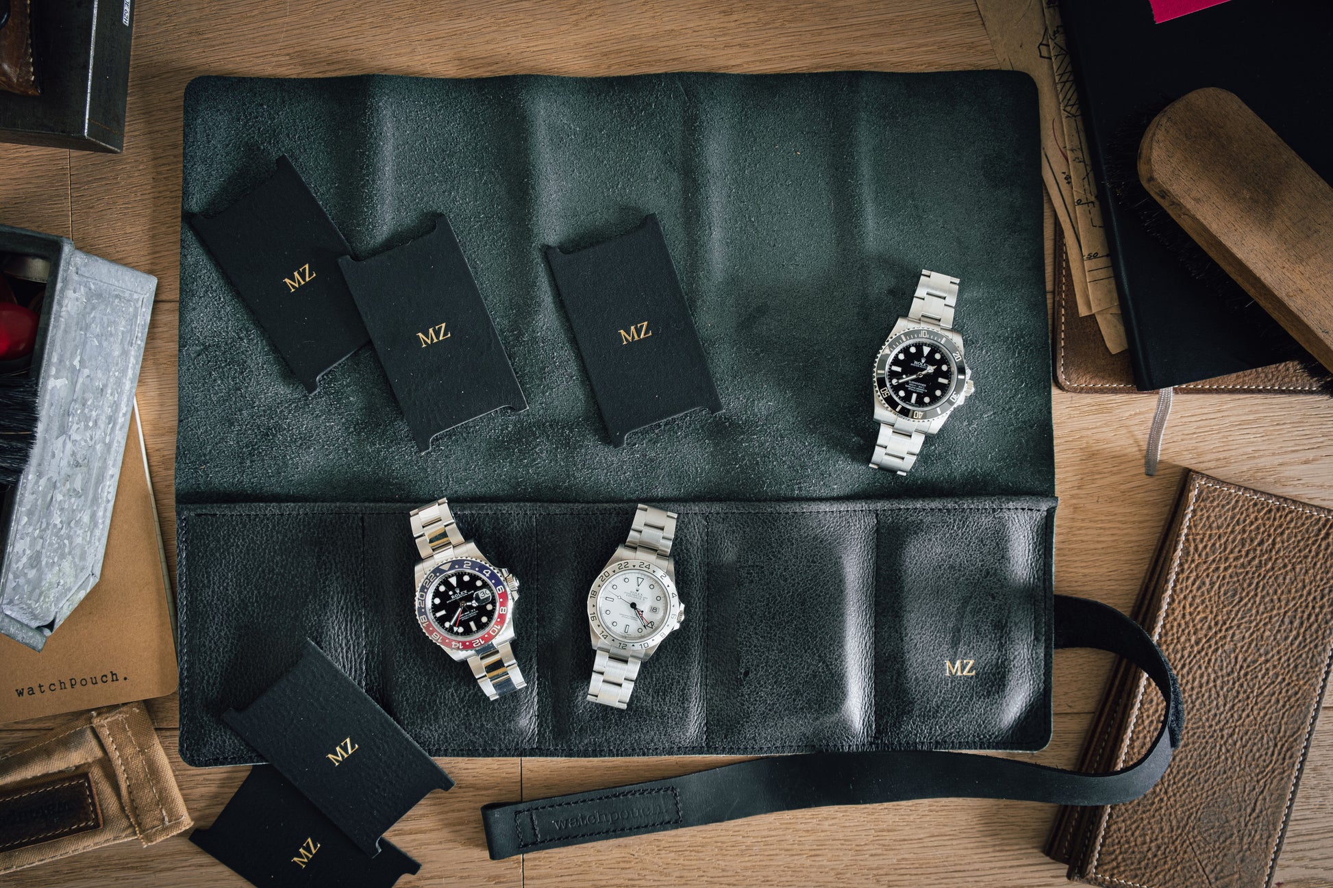 An open black watch roll from the Watchroll collection labeled "MZ" holds three wristwatches with metal straps, meticulously laid out on a wooden surface. Surrounding the personalized watch roll are various leather items, watch pouches, and a stack of books.