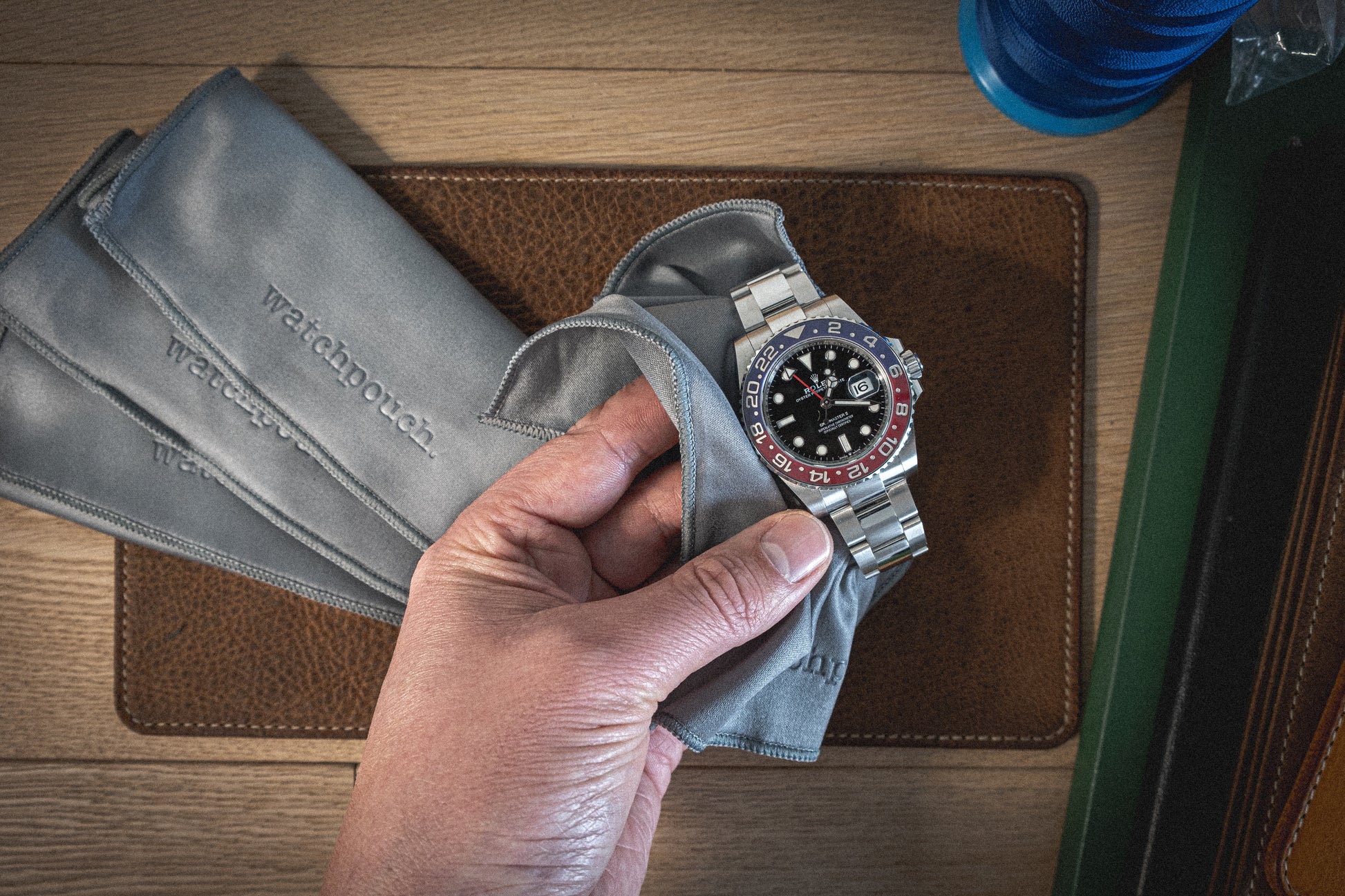 A hand holds a silver wristwatch with a red and black bezel, partially wrapped in one of the soft layered premium pouches (with lining), labeled "Watchbox." The scene includes another premium grey pouch, a brown leather watch mat, and part of a blue cylindrical object to the side.