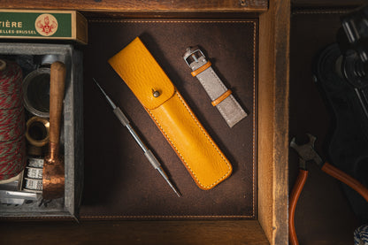 A neatly arranged wooden drawer contains a Toolpouch for springbar-tool. Inside, there is a yellow leather tool pouch holding a springbar tool, an unused grey watch strap essential for watch care, a coil of red string, a matchbox, and various small items. The drawer has a rustic and organized appearance.