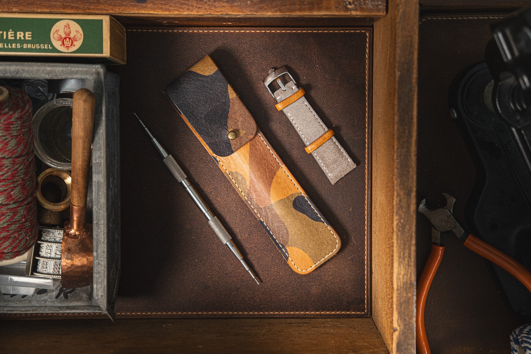 The drawer contains various items: a Toolpouch for Springbar-Tool in a camouflage pattern holding two metallic watch tools, including a springbar tool, alongside a spare watch strap for watch care, a spool of string, a small wooden hammer, a box, and a pair of pliers. The drawer's interior is lined with soft, dark material.