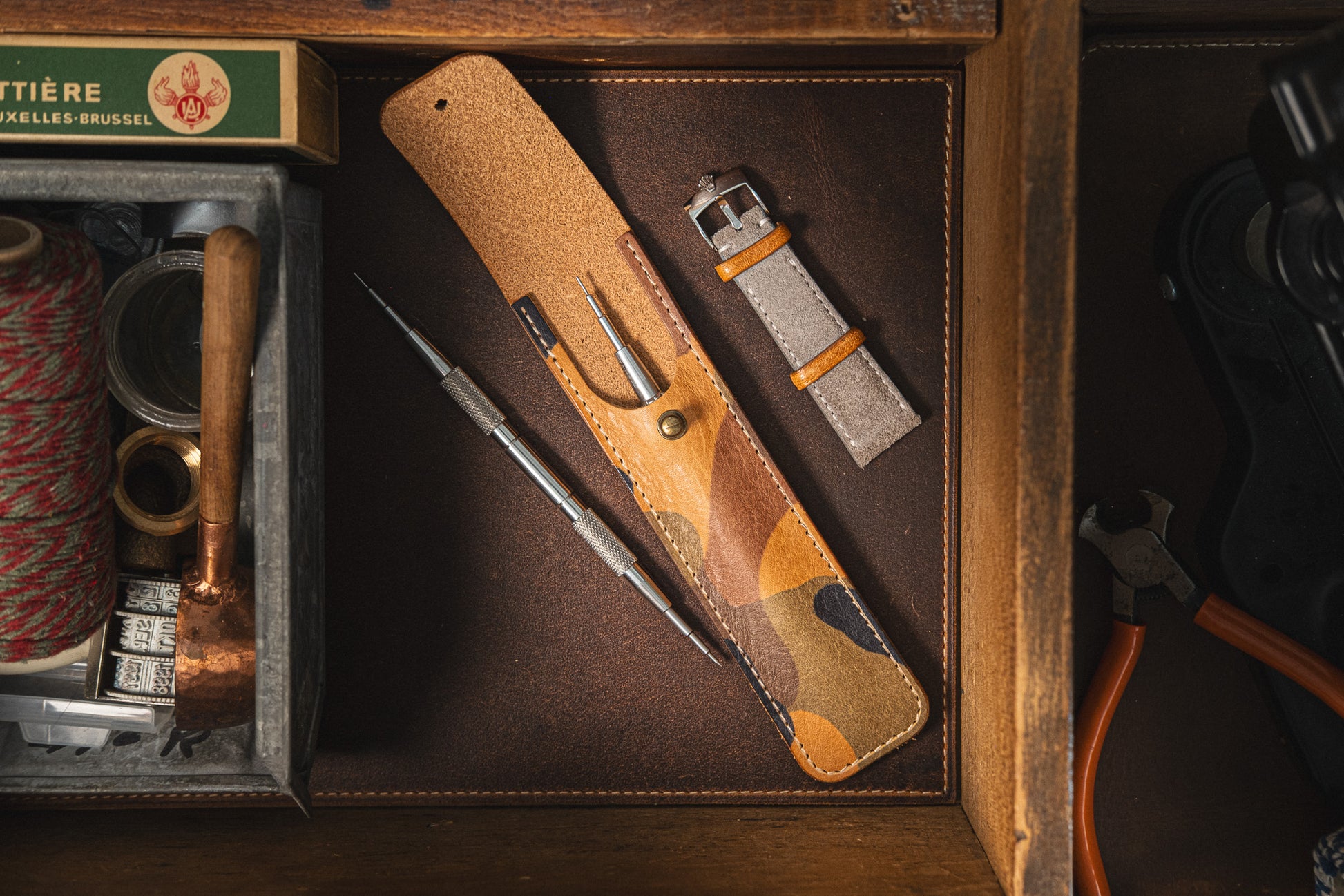A flat lay image of a "Toolpouch for springbar-tool" with a camouflage pattern, containing a springbar tool and a spare watch strap. The pouch is placed on a dark surface surrounded by various tools and supplies such as thread, a small hammer, and a ruler, essential for comprehensive watch care.