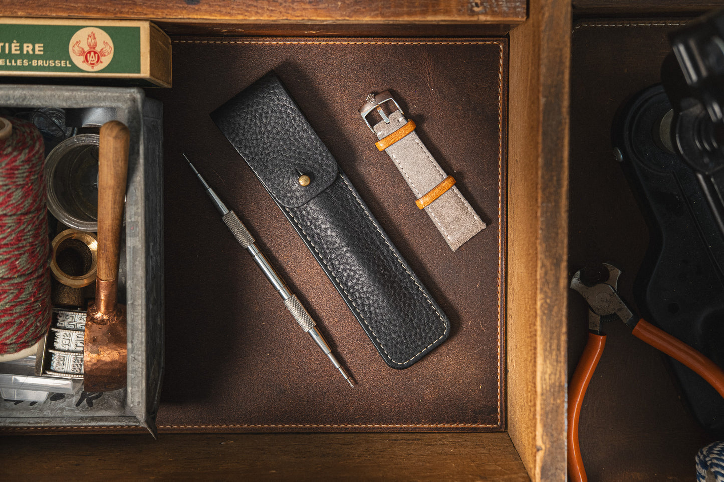 A Toolpouch for the springbar-tool, along with a watch strap and interchangeable heads, is neatly arranged on a leather surface. Nearby, a wooden box holds various tools and supplies for watch care, including a spool of red thread, a small hammer, and other crafting materials.