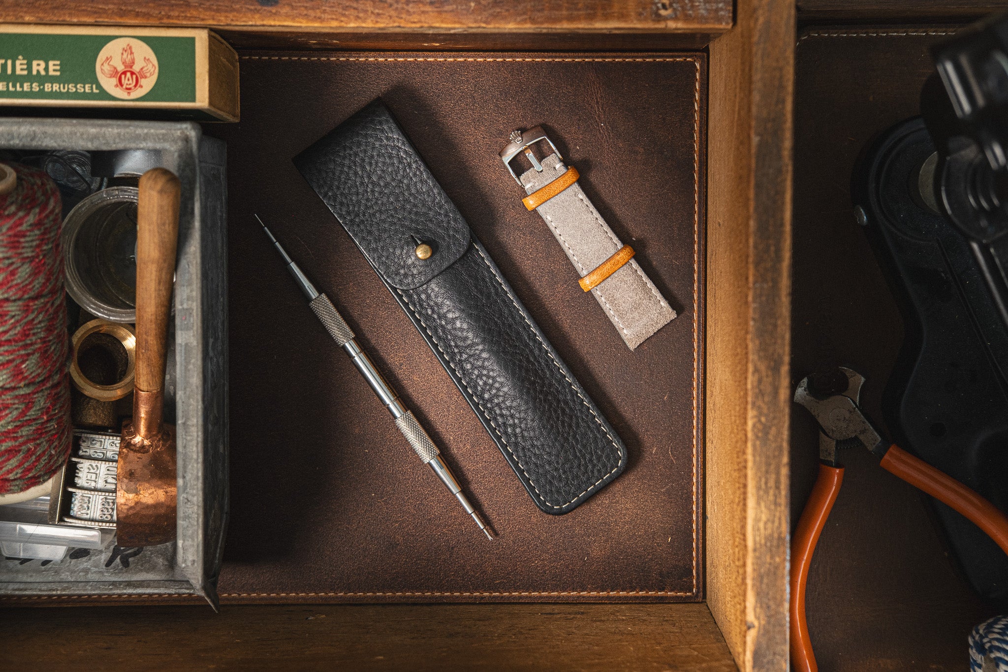 A Toolpouch for the springbar-tool, along with a watch strap and interchangeable heads, is neatly arranged on a leather surface. Nearby, a wooden box holds various tools and supplies for watch care, including a spool of red thread, a small hammer, and other crafting materials.