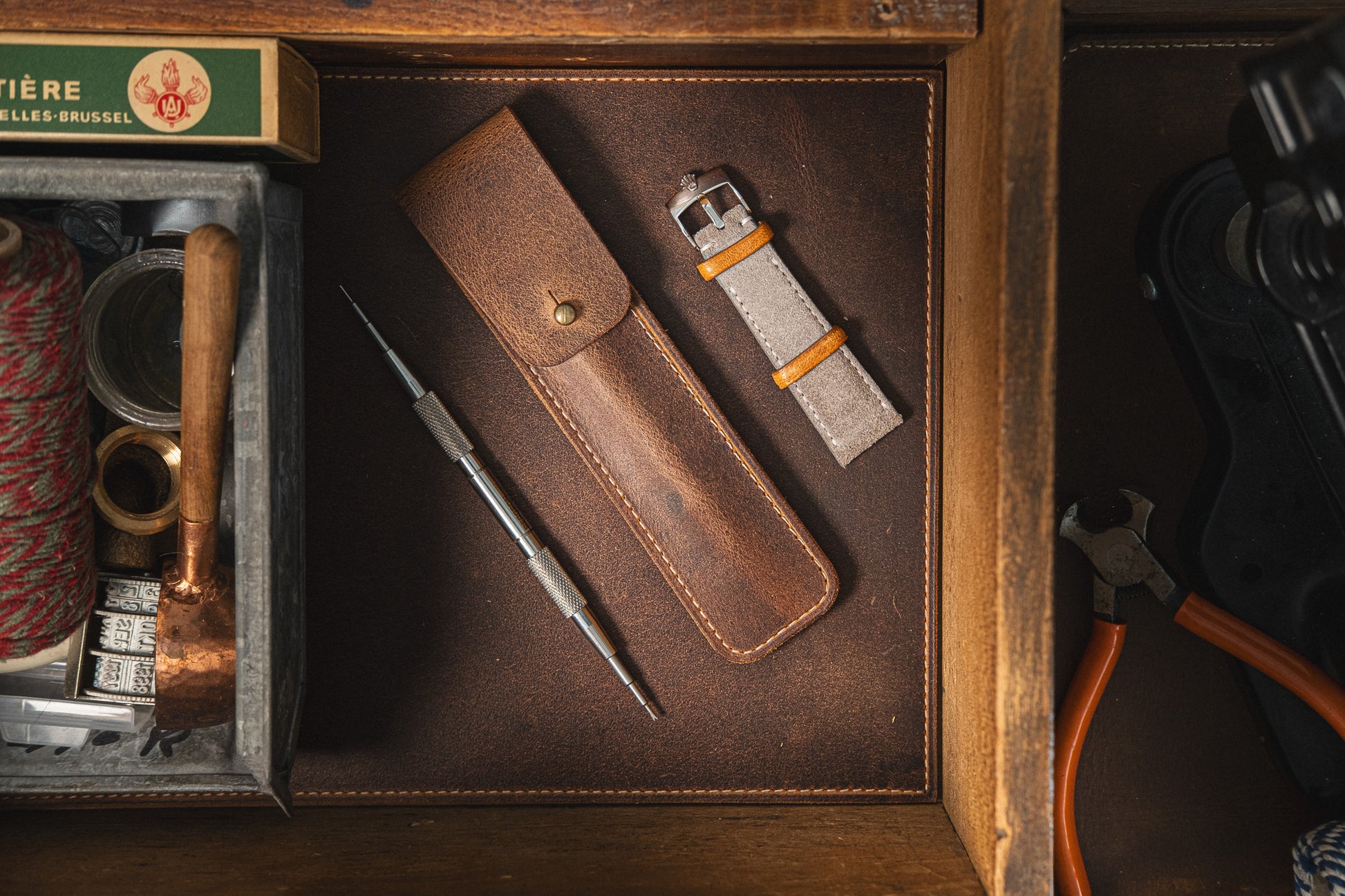 A watch band replacement kit, including a grey fabric watch strap with orange accents, a metal springbar tool, and a leather carrying case, is laid out neatly in a wooden drawer. The background features additional tools and accessories for thorough watch care. Among the items is the Toolpouch for springbar-tool.