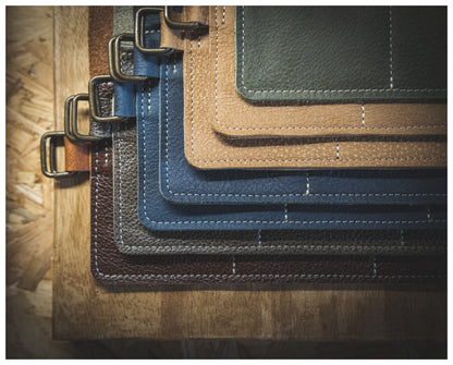 A stack of variously sized rectangular leather swatches from the Watchroll collection, featuring colors such as green, tan, blue, and brown, are neatly arranged on a wooden surface. Each personalized swatch has stitched edges and metal rings on one side, likely for sample display purposes.