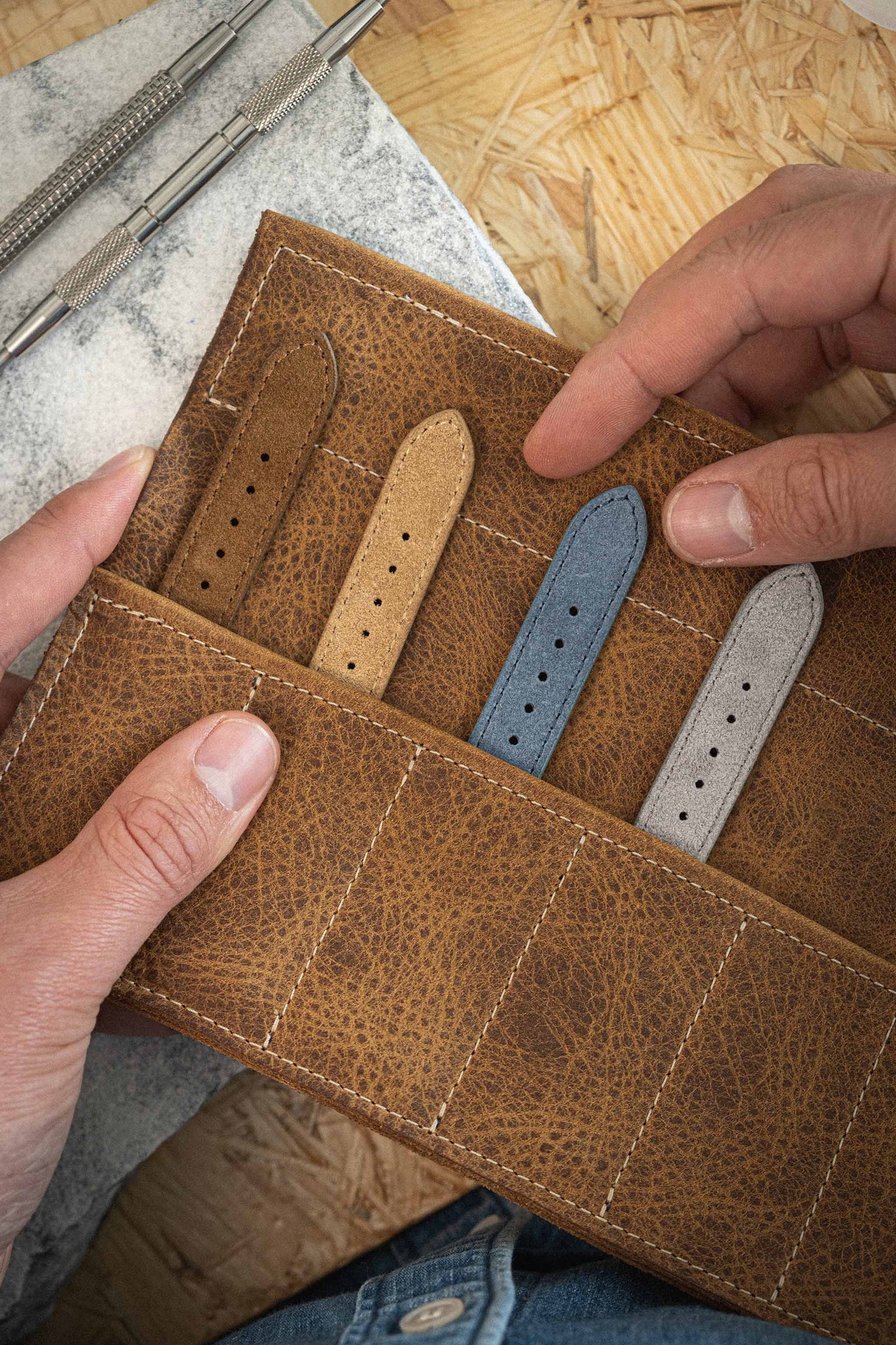 Hands holding a brown leather wallet organizer containing three suede watch straps in tan, blue, and gray. A watch repair tool is visible in the background on a wooden surface. The quick-release spring bar system ensures effortless changes for any occasion.