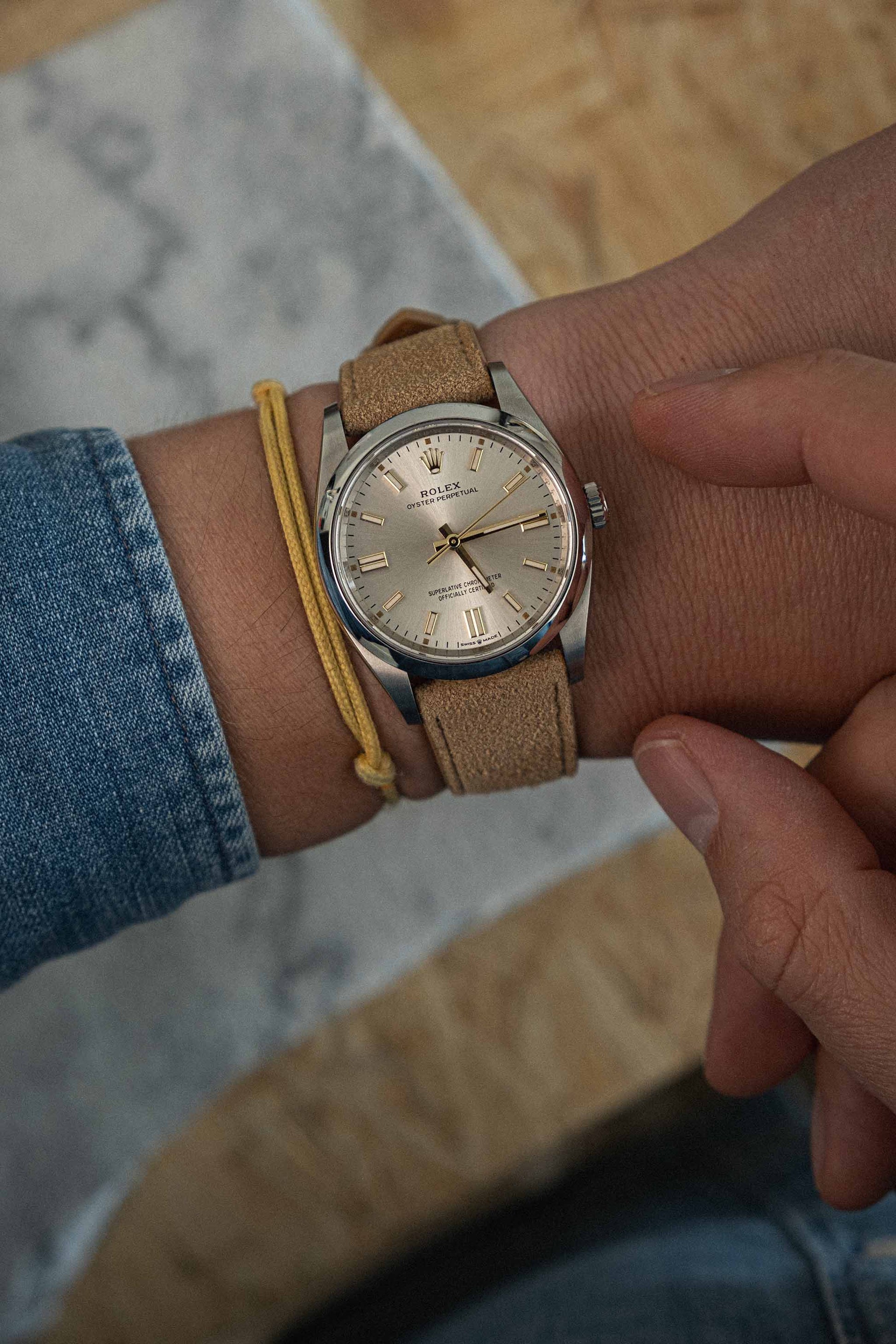 A person wearing a denim jacket and yellow bracelet adjusts a wristwatch with a tan suede strap and a silver watch face displaying the time on their wrist. The background includes wooden and marble surfaces.