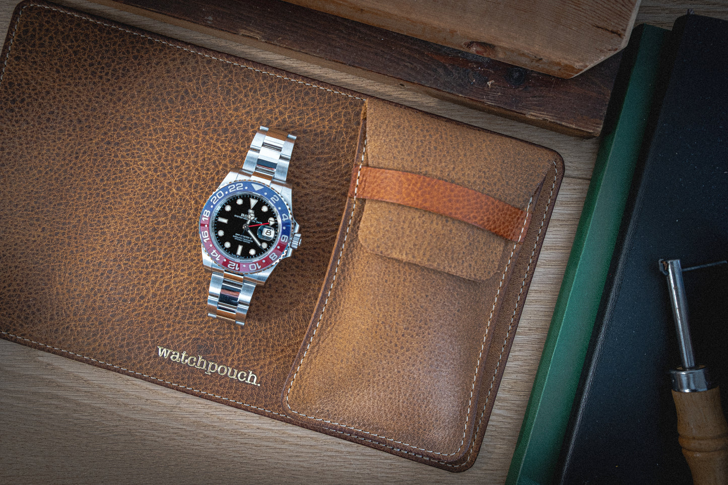 A two-tone red and blue bezel wristwatch with a silver bracelet lies on a Rugged Chestnut leather watch pouch, engraved with "watchpouch" and personalized initials. The pouch is placed on a wooden table with a stack of books in the background, and a corner of a tool is visible on the right.