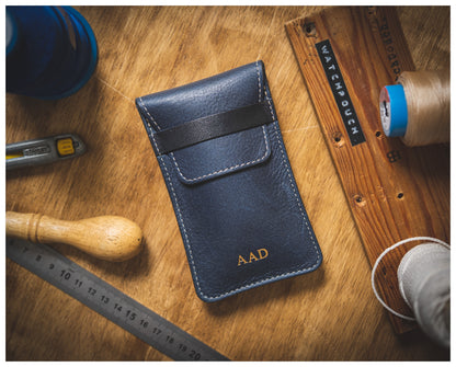 A navy leather pouch embossed with "AAD" sits elegantly on a wooden table surrounded by craft tools—ruler, cutting tool, and spool threads. This piece from the Soft Layered Premium Pouch Collection (with lining), handmade in Antwerp, exemplifies exceptional craftsmanship as a "Watch Pouch.