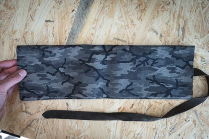 A hand holds a rolled-up watch pouch from the SAMPLE SALE Pebbled veg tan collection featuring a camouflage pattern, with pebbled veg-tan leather initials in lime and cognac strap, all placed on a rustic wooden surface.