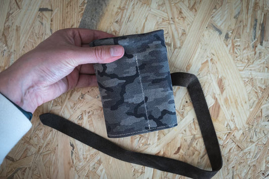 A hand holds a wallet from the SAMPLE SALE Pebbled veg-tan collection, featuring a camouflage design with gray, black, and olive tones and an accompanying cognac strap. The wallet rests on a textured wooden surface.