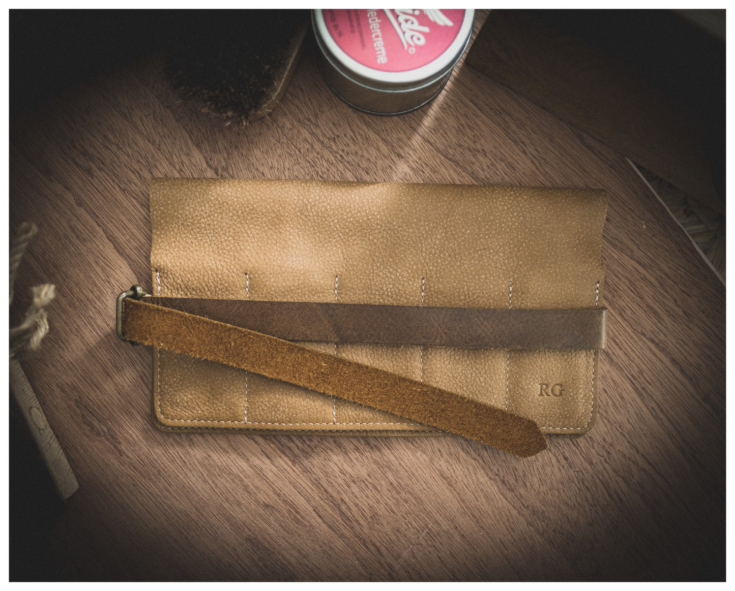 A camel-colored leather wallet with a straproll, featuring 10 pockets, is laid on a wooden surface. The wallet boasts a textured finish and meticulous stitching details. The initials "RG" are embossed in the bottom right corner, adding a touch of personalization. A tin of shoe polish and a brush handle are partially visible in the background.
