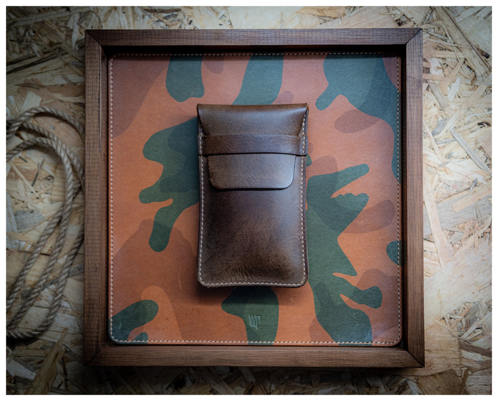 A "vintage brown" watchpouch with a flap closure is placed on a square board covered in camouflage-patterned material. The board is framed with a wooden border and rests on a background of oriented strand board (OSB). A coiled rope is partially visible in the bottom left corner.
