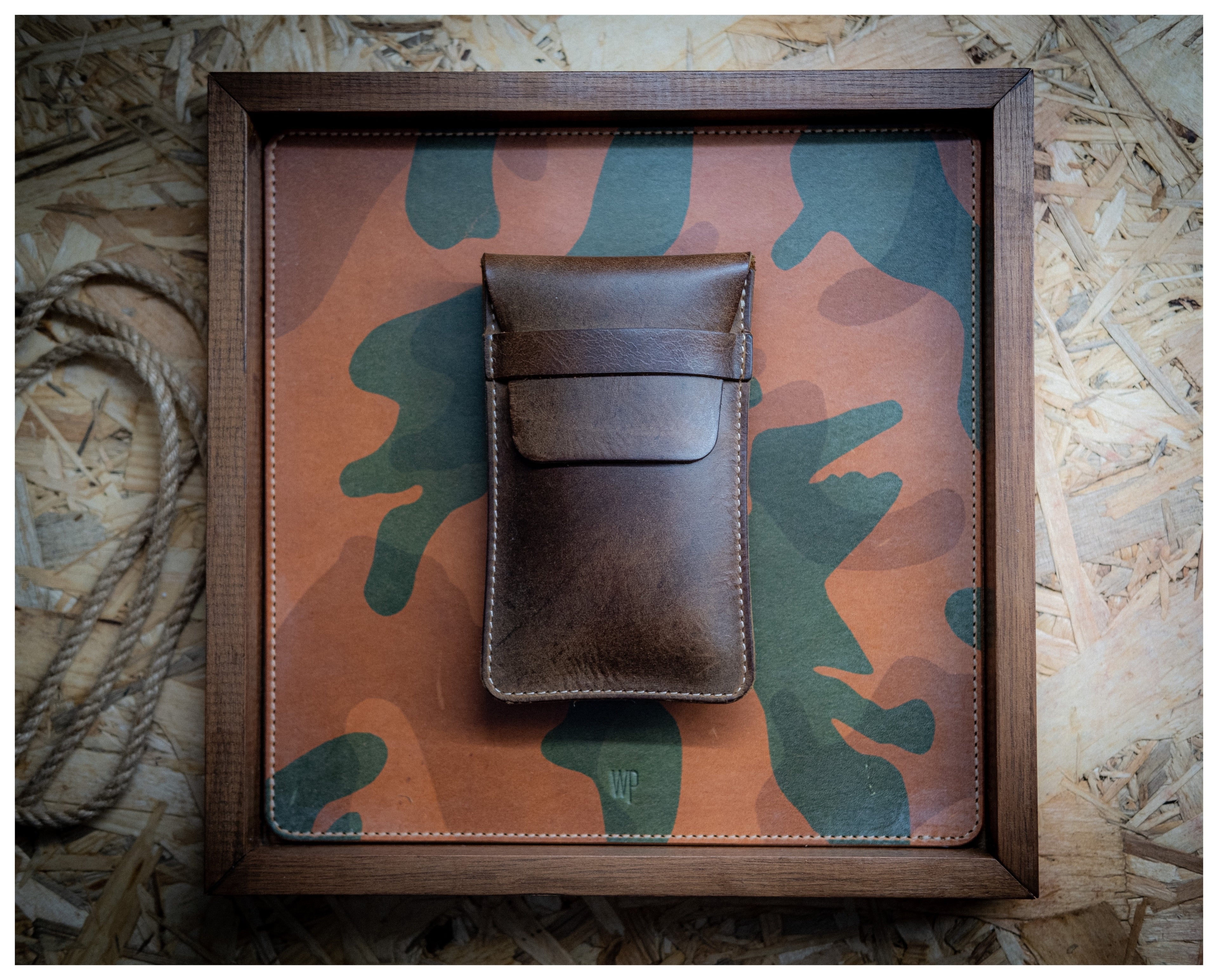 A "vintage brown" watchpouch with a flap closure is placed on a square board covered in camouflage-patterned material. The board is framed with a wooden border and rests on a background of oriented strand board (OSB). A coiled rope is partially visible in the bottom left corner.