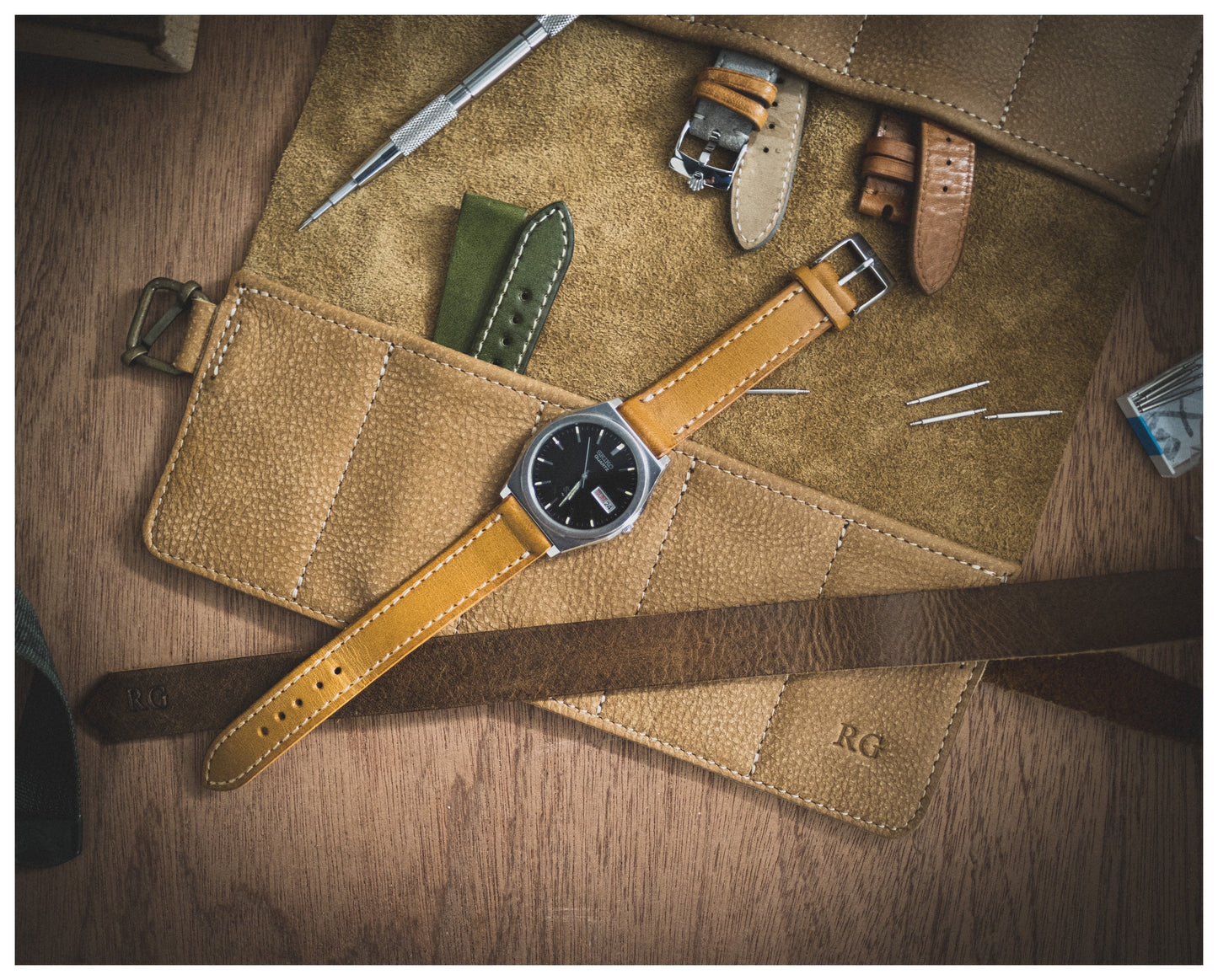 A flat lay image of the straproll “camel”, featuring its soft leather material and 10 pockets, displaying multiple watch straps, a watch with a brown strap, and various watch tools on a wooden surface. The initials "RG" are embossed on the leather roll, adding a touch of personalization.
