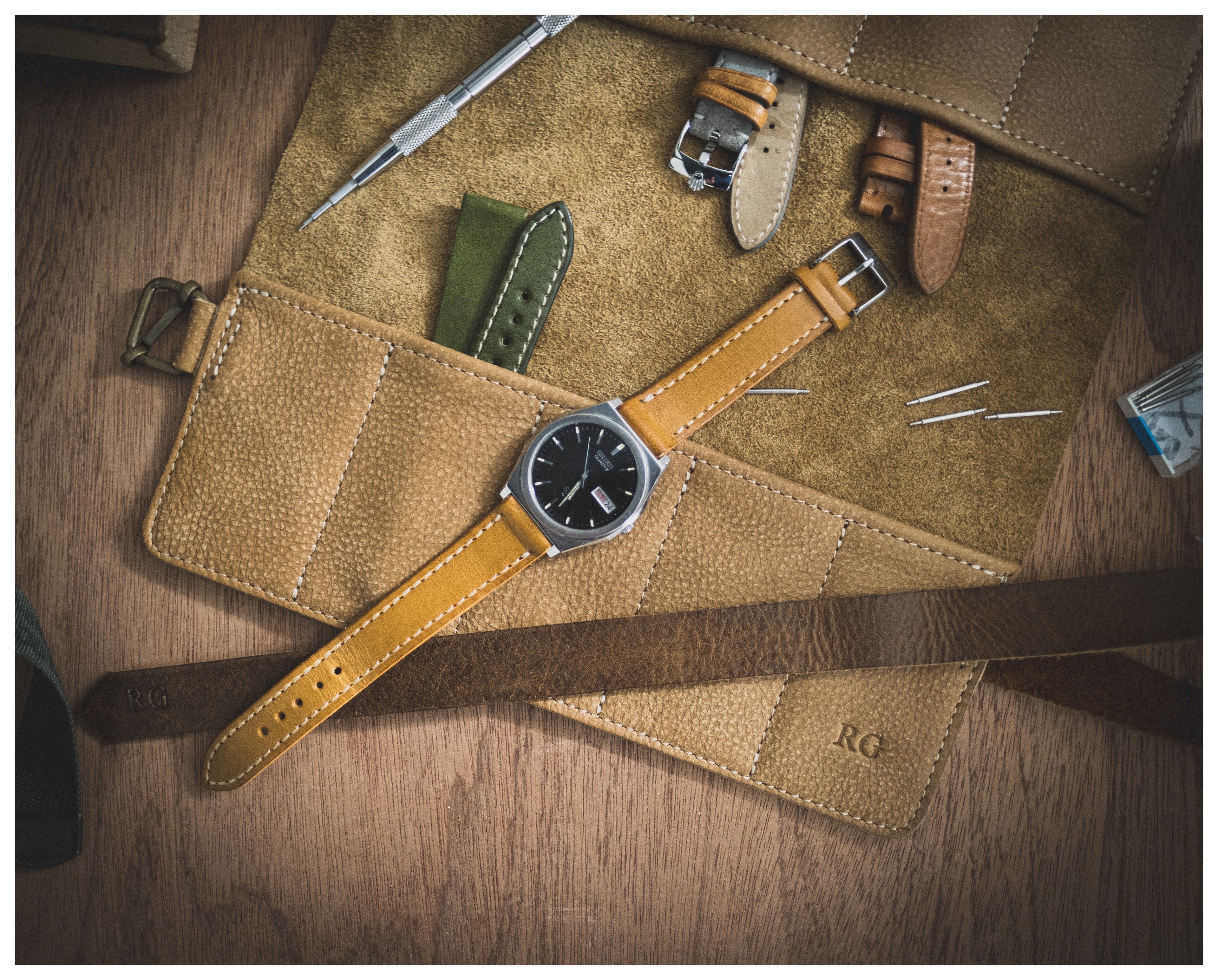 A flat lay image of the straproll “camel”, featuring its soft leather material and 10 pockets, displaying multiple watch straps, a watch with a brown strap, and various watch tools on a wooden surface. The initials "RG" are embossed on the leather roll, adding a touch of personalization.