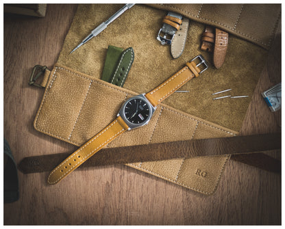 A flat lay image of the straproll “camel”, featuring its soft leather material and 10 pockets, displaying multiple watch straps, a watch with a brown strap, and various watch tools on a wooden surface. The initials "RG" are embossed on the leather roll, adding a touch of personalization.
