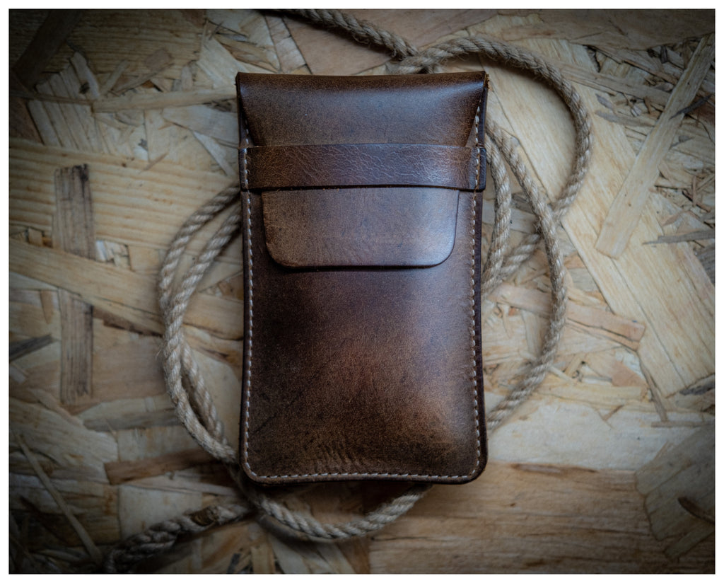 A "vintage brown" leather watchpouch with a flap closure rests on a wooden surface. A thick, coiled rope is partially visible beneath the pouch, creating a rustic backdrop. The patina leather appears worn, adding an even more authentic vintage aesthetic to the image.