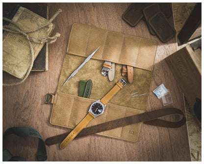 A neatly arranged straproll "camel" with 10 pockets holds a pair of watch straps, a watch tool, and several small accessories. A personalized yellow leather-banded watch lies on top. Nearby are a soft leather strap holder, book, and other small items on a wooden surface.